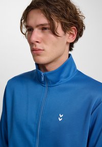Blå zip-jacka i slätt tyg, med hög krage och en liten vit logotyp med tre chevroner på vänster sida.