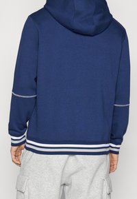 Felpa con cappuccio blu navy con righe bianche e grigie a coste sui polsini e sull'orlo. Realizzata in materiale morbido, presenta una tasca a marsupio.