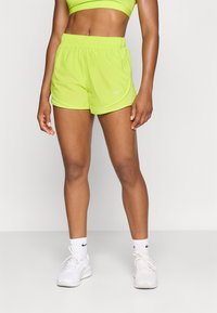 Neonski zeleni atletski šorts s elastičnom trakom u struku i suptilnim Nike logom, uparen s bijelim tenisicama i čarapama do gležnja. Glatka tekstura tkanine.