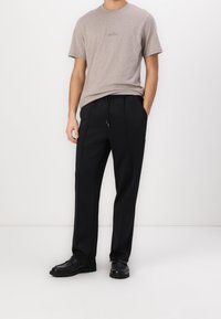 Graues Herren-T-Shirt mit Rundhalsausschnitt und dezentem Logo, kombiniert mit schwarzen locker sitzenden Hosen mit Kordelzug und seitlichen Nähten. Schwarze Schuhe.