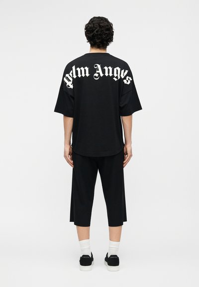Persoană cu păr creț și întunecat stând cu fața spre departe, purtând o cămașă neagră supradimensionată cu textul „Palm Angels” pe spate, pantaloni negri largi, croppați, și adidași.