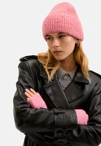 Rosa ribbad stickad mössa prydd med små dekorativa nitar, tillsammans med en svart läderjacka och rosa stickade ärmslut, mot en neutral bakgrund.