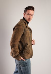 Brun læderjakke med en slidt tekstur, lynlås foran og sidelommer, båret over en sort turtle neck og kombineret med blå jeans.