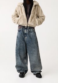 Giacca corta in pelliccia sintetica beige, top grigio, jeans denim blu oversize, cintura nera con fibbia argentata e stivali neri chunky.