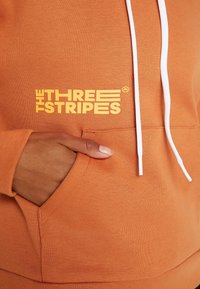 Main reposant dans la poche avant d’un sweat à capuche orange avec des cordons blancs et un texte jaune indiquant « THE THREE STRIPES ».