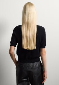 Maglione nero a maglia con maniche corte, abbinato a pantaloni di pelle neri a vita alta. I capelli lunghi e lisci sono biondi. Sfondo minimalista.
