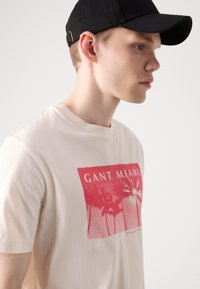 Krémové tričko s krátkým rukávem a červenou grafikou "GANT MIAMI" s palmami. Hladká bavlněná látka, kulatý výstřih, volný střih.