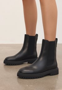 Svarta läderankelboots med kraftiga gummisulor, bärs på bara ben och står på ett polerat betonggolv.