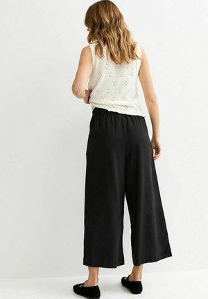 Donna vista da dietro che indossa un top bianco senza maniche in maglia, pantaloni neri a gamba larga e corta, e scarpe nere slip-on su sfondo bianco.