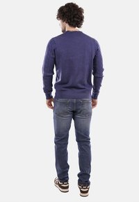 Maglione viola lavorato a maglia con maniche texturizzate, abbinato a jeans blu e sneakers marroni, visto da dietro su uno sfondo semplice.