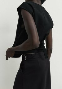 Top noir sans manches au design structuré, fabriqué dans un matériau texturé. Possède des coutures courbes et un lien à l'arrière pour l'ajustement.