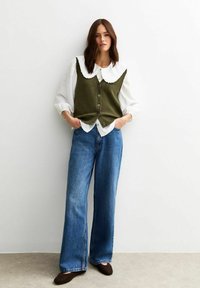 Gilet tricoté vert olive avec un col en dentelle blanc, associé à une chemise blanche ample et un jean bleu en denim large. Des chaussures marron complètent le look.
