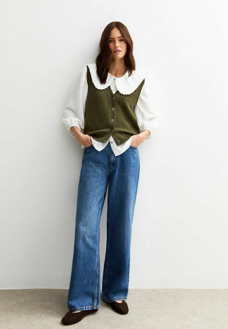 Gilet tricoté vert olive avec un col en dentelle blanc, associé à une chemise blanche ample et un jean bleu en denim large. Des chaussures marron complètent le look.
