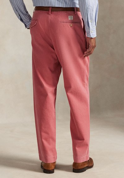 Ροζ βαμβακερό παντελόνι με άνετη εφαρμογή, διαθέτει διπλές πίσω τσέπες και μια διακριτική ετικέτα του brand πάνω από τη δεξιά τσέπη. Φορεμένο με καφέ παπούτσια.