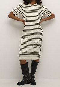 Robe en maille rayée crème et noir, manches courtes, silhouette ajustée. Associée à de hautes bottes en cuir noir avec des accents à boucle.