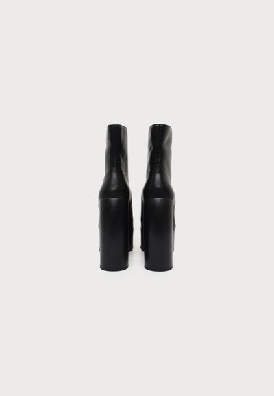 Paire de bottes noires à plateforme avec de hauts talons épais vue de dos sur un fond blanc uni.