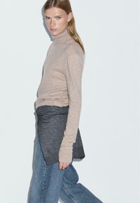 Maglione beige a collo alto con una texture morbida sovrapposto a una maglietta grigia. Jeans a gamba larga di un blu chiaro strappati completano l'outfit.