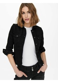 Veste en denim noire avec fermeture à boutons, deux poches poitrine et manches retroussées, associée à une chemise blanche et un pantalon noir.