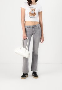 Camiseta blanca corta con un gráfico de oso de peluche, jeans acampanados grises y un bolso blanco. Zapatillas negras con rayas blancas completan el atuendo.