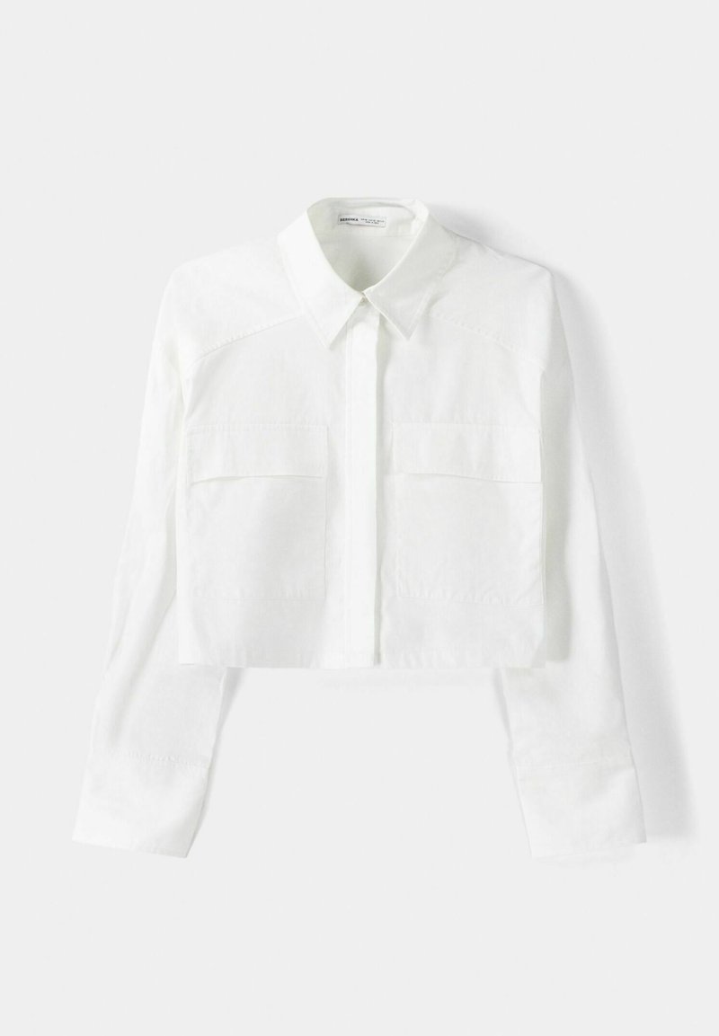 Camicia bianca tagliata realizzata in tessuto testurizzato, con colletto a punta, chiusura a bottoni e due tasche sul petto. Maniche lunghe con polsini.