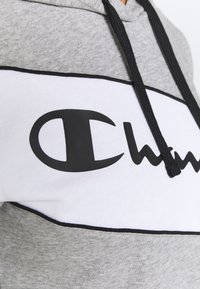 Felpa grigia con cappuccio caratterizzata da un pannello bianco con un logo nero prominente, dettagli neri e coulisse, realizzata in un morbido tessuto misto cotone.