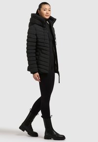 Giacca imbottita nera con cappuccio, caratterizzata da un design trapuntato, chiusura frontale con zip e tasche laterali. Abbinata a leggings neri e stivaletti alla caviglia.
