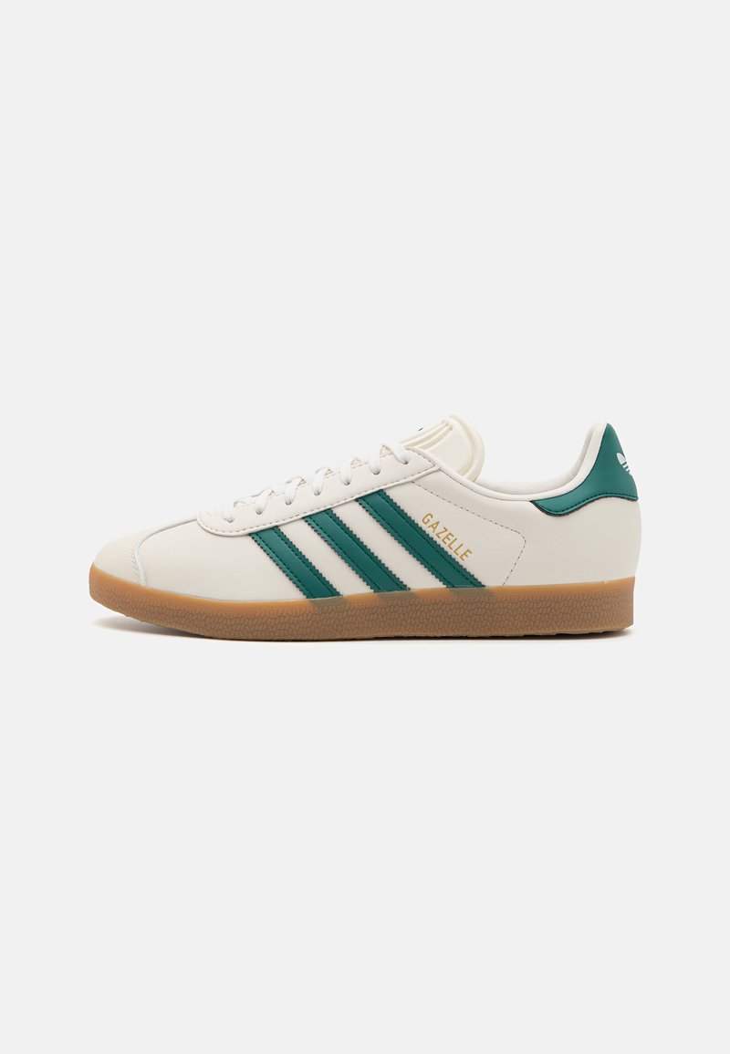 Baltie sneakeri ar zaļām svītrām, ar gumijas zoli, apaļu purngalu un tekstūrētu ādas materiālu. "Gazelle" logo zeltā.