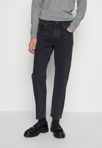 Sorte denim jeans med rett snitt, som har en klassisk fem-lomme design og subtil søm. Kombinert med en grå genser og sorte sko.