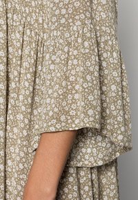 Robe à motif avec des manches courtes et évasées. Tissu vert olive avec de petites fleurs blanches et une apparence texturée. Détail plissé qui accentue le design.