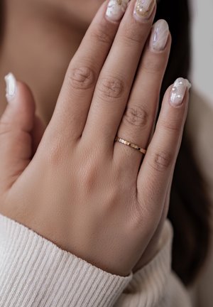 Goldener Ring mit strukturierten Segmenten an einer Hand, mit Nägeln, die einen hellen Nagellack und Verzierungen aufweisen. Leichtes Stoffmanschette sichtbar.