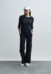 Svart langermet skjorte med tekst, svarte cargobukser med glidelåser og lommer, kombinert med hvite joggesko og en hvit caps med striper.