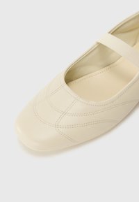 Beige skinnballerina med firkantet tå, med et sydd mønster og en elastisk stropp. Glatt tekstur og minimalistisk design.