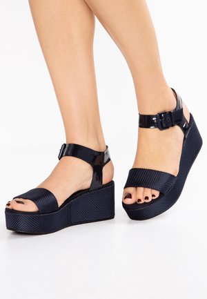 Pieds portant des sandales à plateforme compensée noires avec des brides à la cheville et des orteils ouverts, montrant des ongles de pieds peints en foncé sur fond blanc.