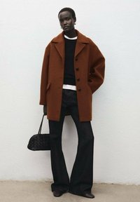 Bruine wollen jas met grote knopen, zwarte trui en uitlopende donkere spijkerbroek. Houdt een zwarte gevlochten handtas vast. Simpele achtergrond.