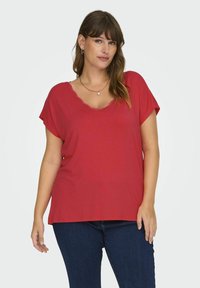 Červené tričko s krátkým rukávem a vlnitým výstřihem, volného střihu, vyrobené z měkkého materiálu, v kombinaci s tmavomodrými džíny.