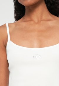 Λευκό ριμπ τοπάκι με λεπτές τιράντες και μικρό μεταλλικό λογότυπο στο κέντρο του στήθους. Το ύφασμα έχει λεία υφή.