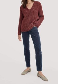 Femme portant un pull col V côtelé bordeaux, un pantalon en velours côtelé bleu marine, et des ballerines beiges pointues, debout avec une main dans la poche.