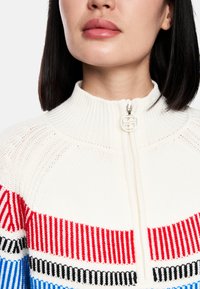 Knit zip-up sweater in cream with red, black, and blue horizontal stripes. Features a high collar and a circular logo zipper pull.