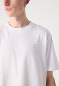 Bílá bavlněná tričko s krátkým rukávem, na levé straně hrudi s světle modrým logem "ck". Jednoduchý, klasický design a měkká textura.