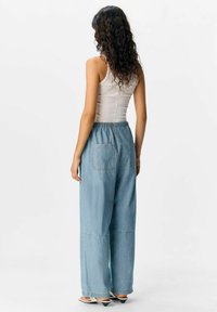 Pantaloni in denim blu chiaro a gamba larga con vita regolabile, dotati di due tasche posteriori e cuciture a contrasto, abbinati a una canotta aderente.