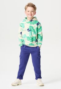 Grüne und beige Kapuzenjacke mit Blattmuster, kombiniert mit lila Cargo-Hosen. Weiße Sneaker mit strukturiertem Detail runden das Outfit ab.