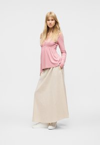 Νεαρή γυναίκα στέκεται με τα χέρια στις τσέπες, φορώντας ροζ μακρυμάνικη μπλούζα, μπεζ μακριά φούστα και λευκά αθλητικά παπούτσια σε απλό φόντο.