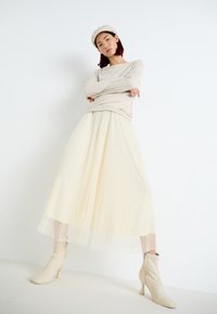 Beige langærmet top i kombination med en plisseret cremefarvet nederdel. Modellen har lyse ankelstøvler på med lynlåsdetalje. Enkel, elegant design.