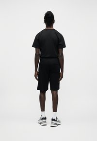 Άνδρας που στέκεται με την πλάτη προς τα εμπρός, φορώντας μαύρο μπλουζάκι, μαύρο σορτς, λευκές κάλτσες και μαύρα-άσπρα αθλητικά παπούτσια με λευκό φόντο.