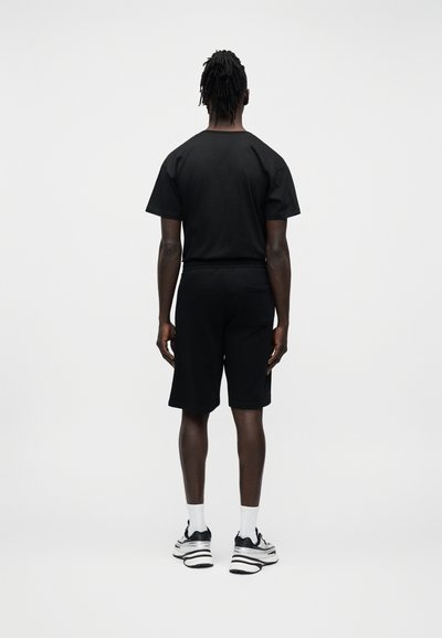 Homme de dos portant un t-shirt noir, un short noir, des chaussettes blanches et des baskets noires et blanches, sur fond blanc.