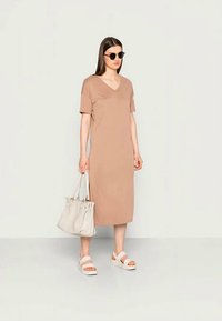 Vestuário castanho até ao joelho com mangas curtas e decote em V. Acompanhado por sandálias brancas de plataforma e uma mala cinza-claro. Óculos de sol usados.