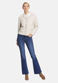 Hellbeige Tweed-Jacke mit Rundhalskragen, zwei vorderen Taschen, kombiniert mit blauen Schlagjeans und hellbraunen spitzen Pumps.