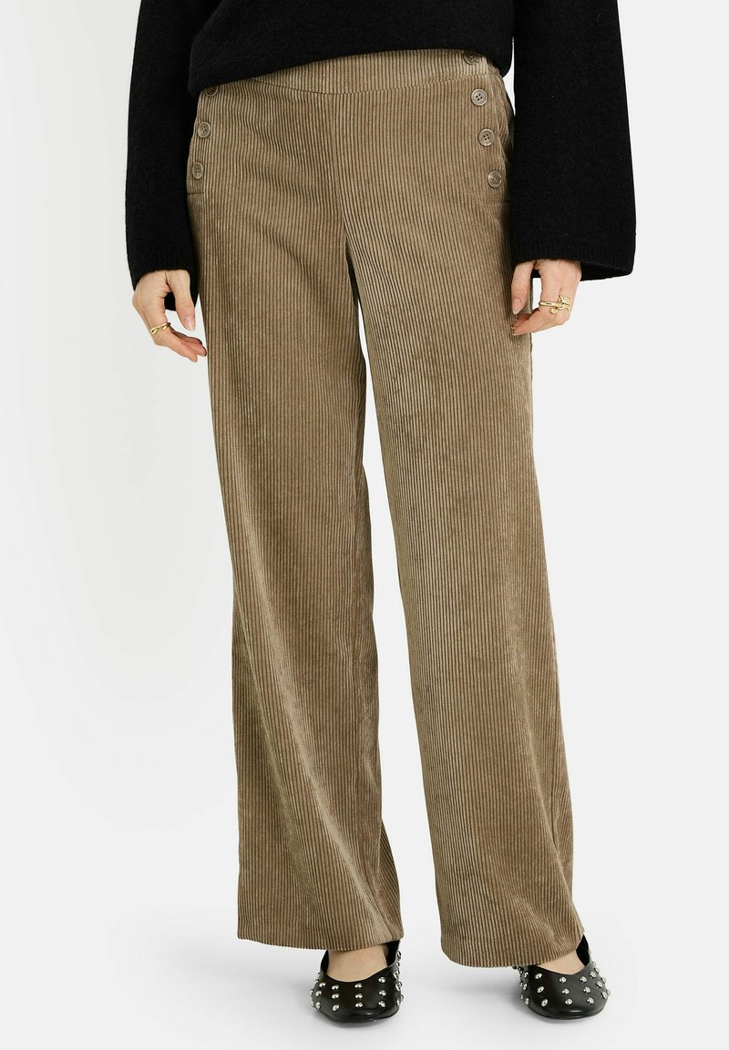 Pantalones anchos de pana marrón con rayas verticales, con cuatro botones decorativos en la cintura y una textura suave. Combinados con zapatos planos negros.