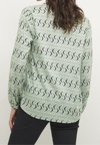 Vaaleanvihreä pusero, jossa on musta abstrakti aaltoaihe, pitkät hihat joustavilla resoreilla ja pyöristetty helma.