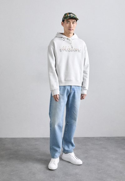 Sweat à capuche gris clair avec "Missoni" en lettres décoratives, associé à un jean droit bleu clair et des baskets blanches. Casquette avec un motif coloré.